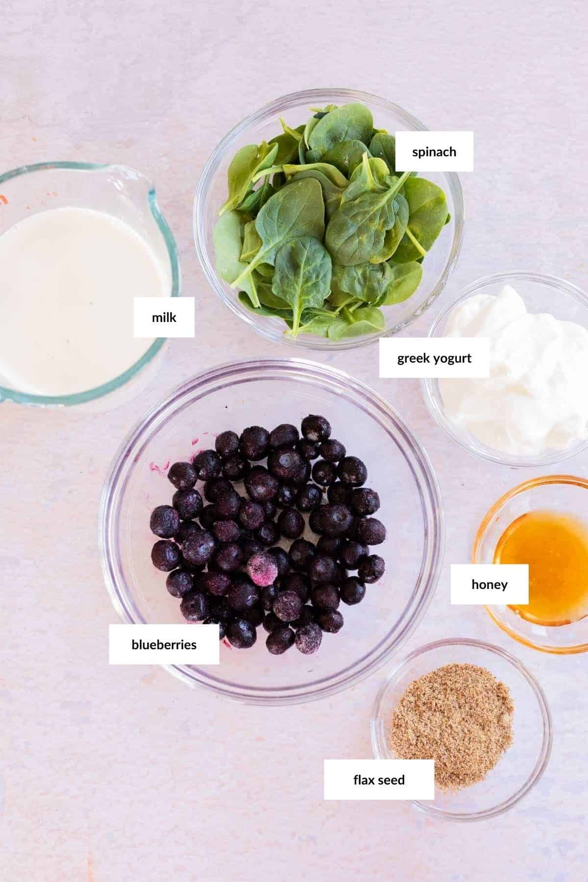 Displayed on the table are the labeled ingredients for a delicious blueberry smoothie: milk, spinach, Greek yogurt, blueberries, honey, and flax seed.