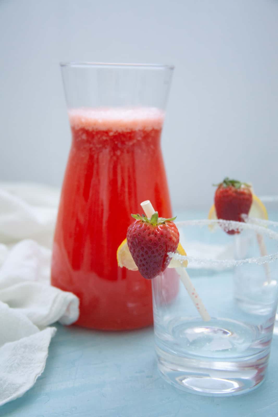 If you're looking for a perfectly easy to make mocktail, this strawberry lemonade mocktail is it! It's so easy to make and enjoy whether you are at a party or just at home enjoying the weather outside! a white kitchen towel with red strawberry lemonade mocktail in a pitcher with two empty glasses garnished with strawberries