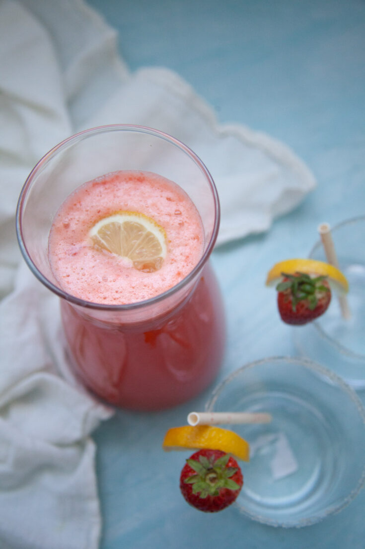 a pitcher of strawberry lemonade mocktail with two empty glasses on a blue background