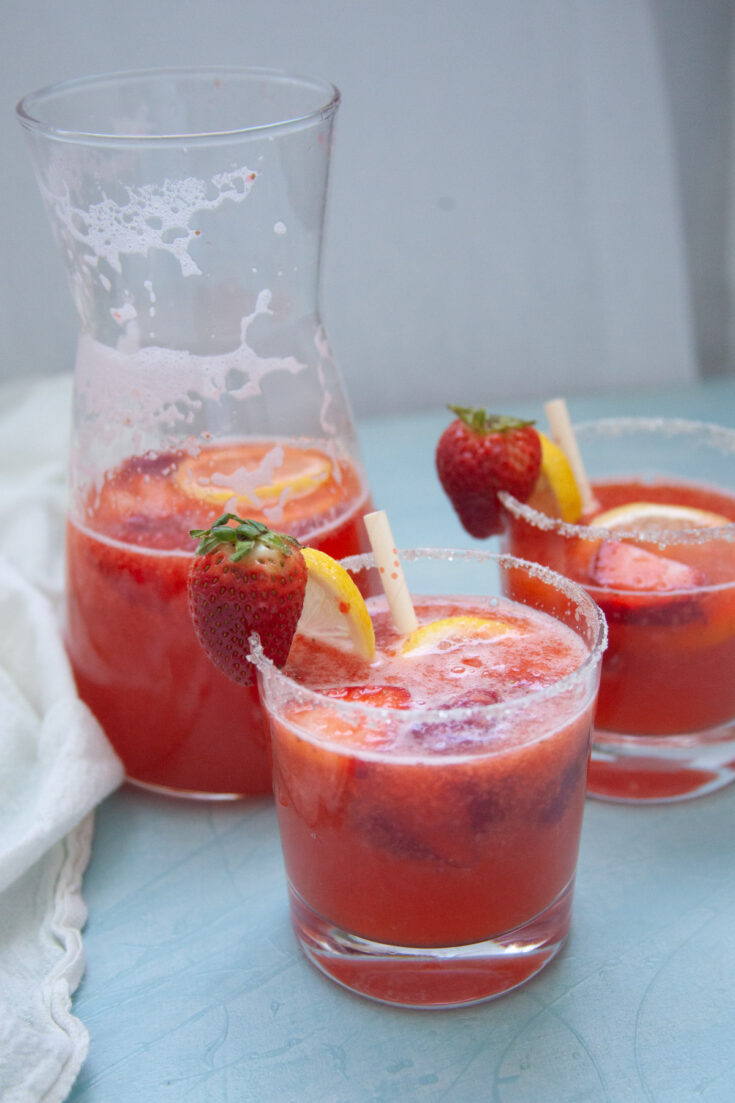 two strawberry lemonade mocktails on a blue background with a pitcher