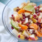 a glass bowl of apples, pears, pomegranate seeds, lime wedges and orange wedges in a thanksgiving fruit salad on a blue background