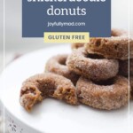 A stack of pumpkin snickerdoodle donuts, one with a bite taken out, on a white plate with a label indicating they are gluten-free and from joyfullymad.com, next to a dish of lemon pepper chicken thighs.