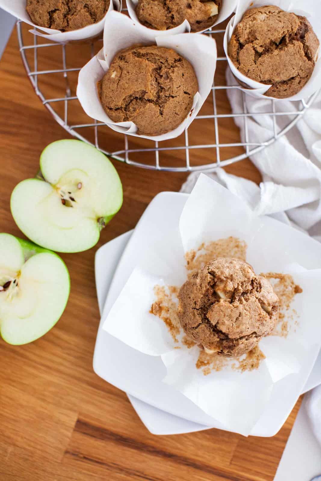 Apple cinnamon muffins wrapped in parchment paper on a wire rack with apple halves nearby.
