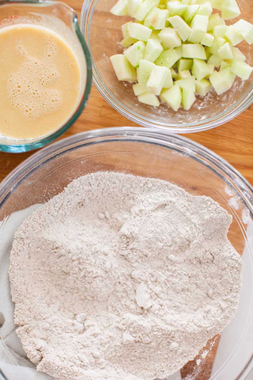 A clear mixing bowl of dry ingredients, with smaller bowls of diced apple and wet ingredients.