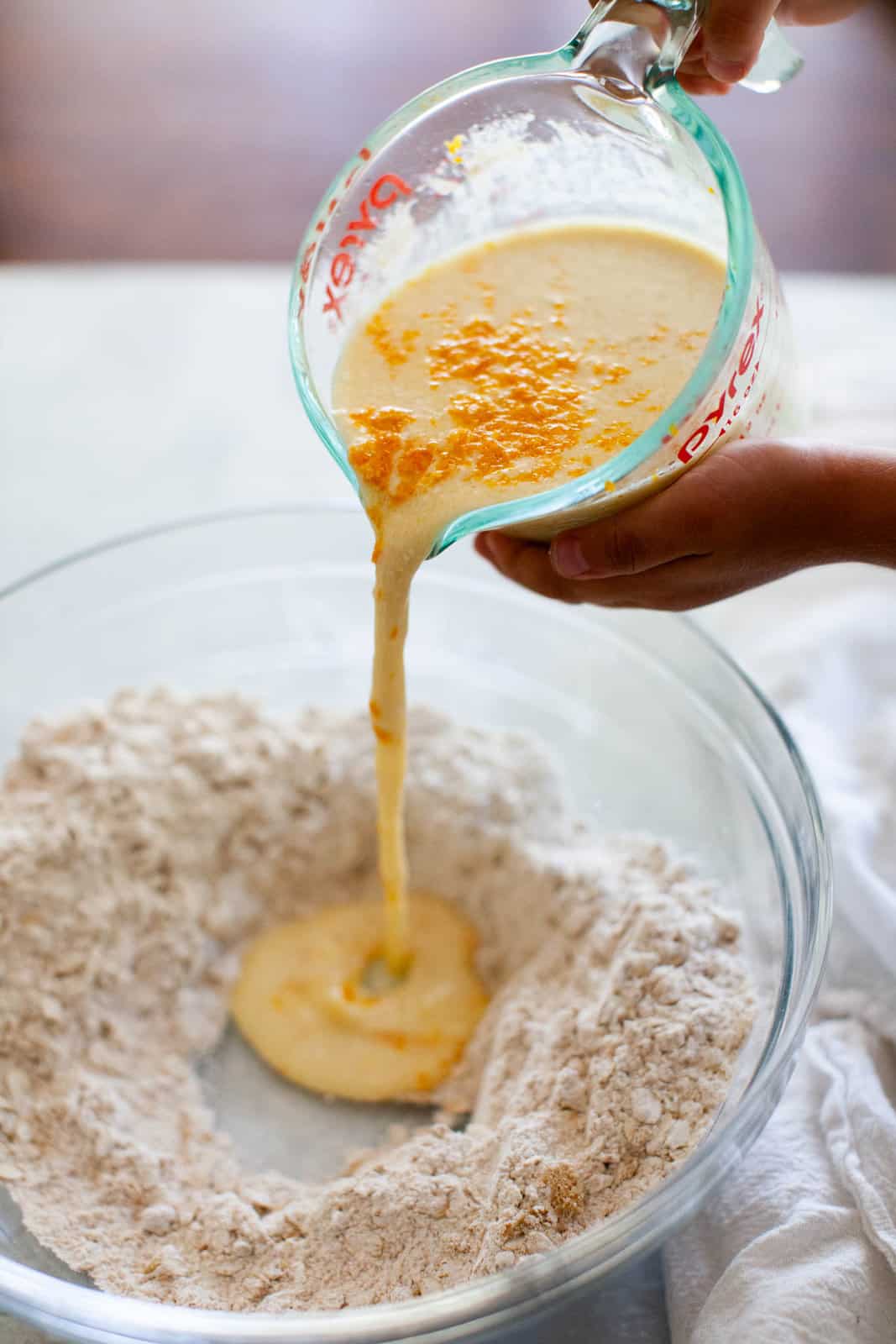 Pouring wet mixture from a glass measuring cup into a bowl of flour, sugar, and rolled oats.
