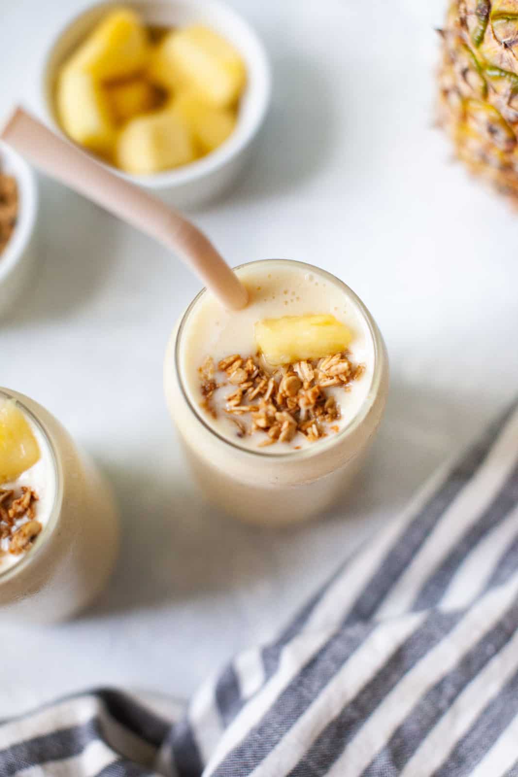 Overhead view of a pineapple drink garnished with fresh pineapple chunks, granola, and a straw.