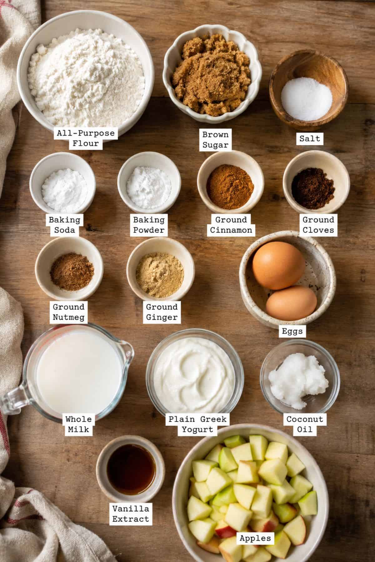 Top-down view of labeled baking ingredients in bowls and cups on a wooden surface, including flour, sugars, spices, eggs, milk, yogurt, coconut oil, vanilla extract, and chopped apples in individual bowls.