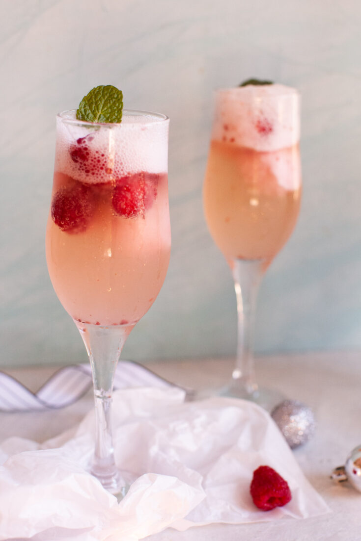 a raspberry lies next to a champagne flute with a pink champagne cocktail.