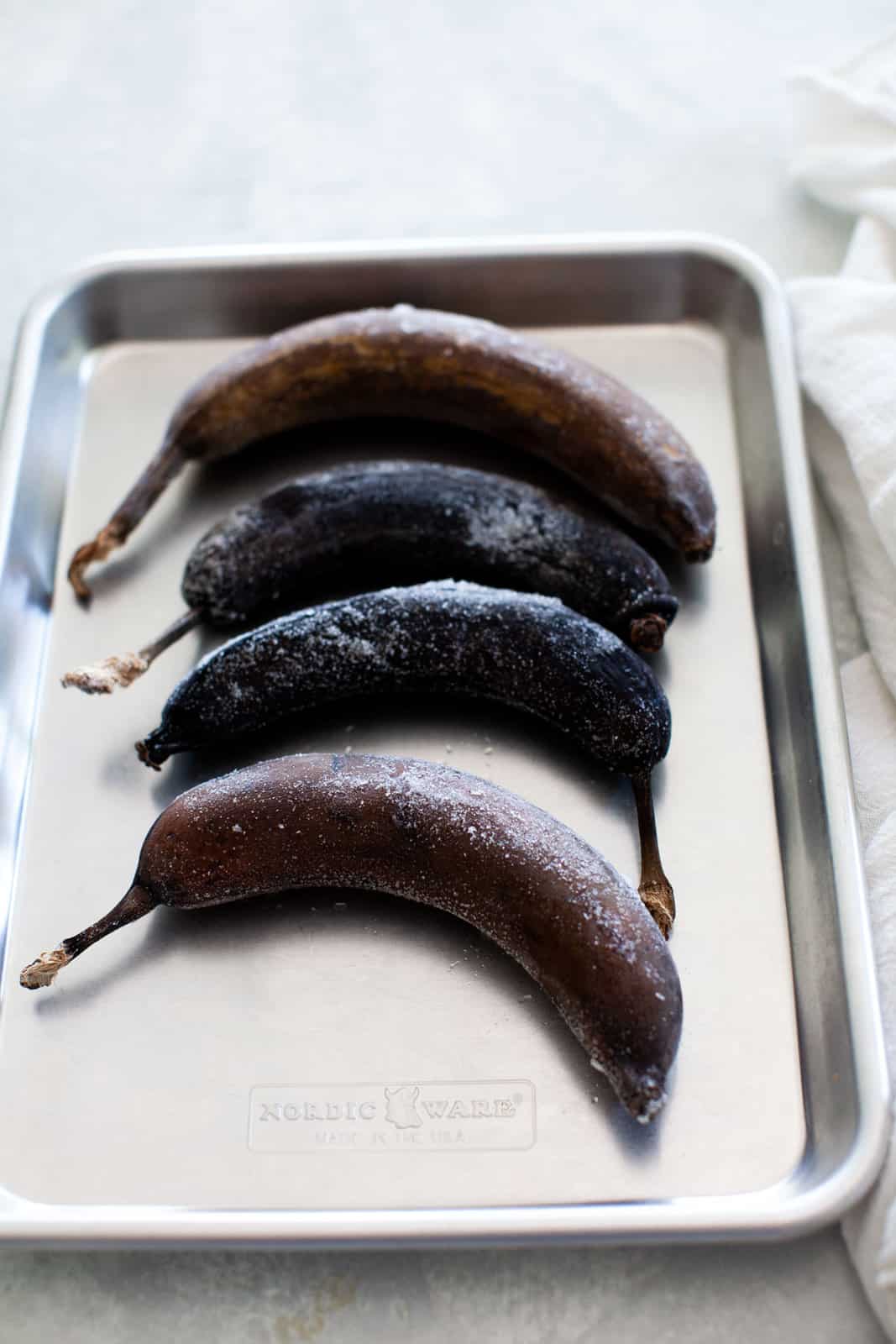 Four whole frozen bananas with dark peels are arranged in a row on a metal baking tray.