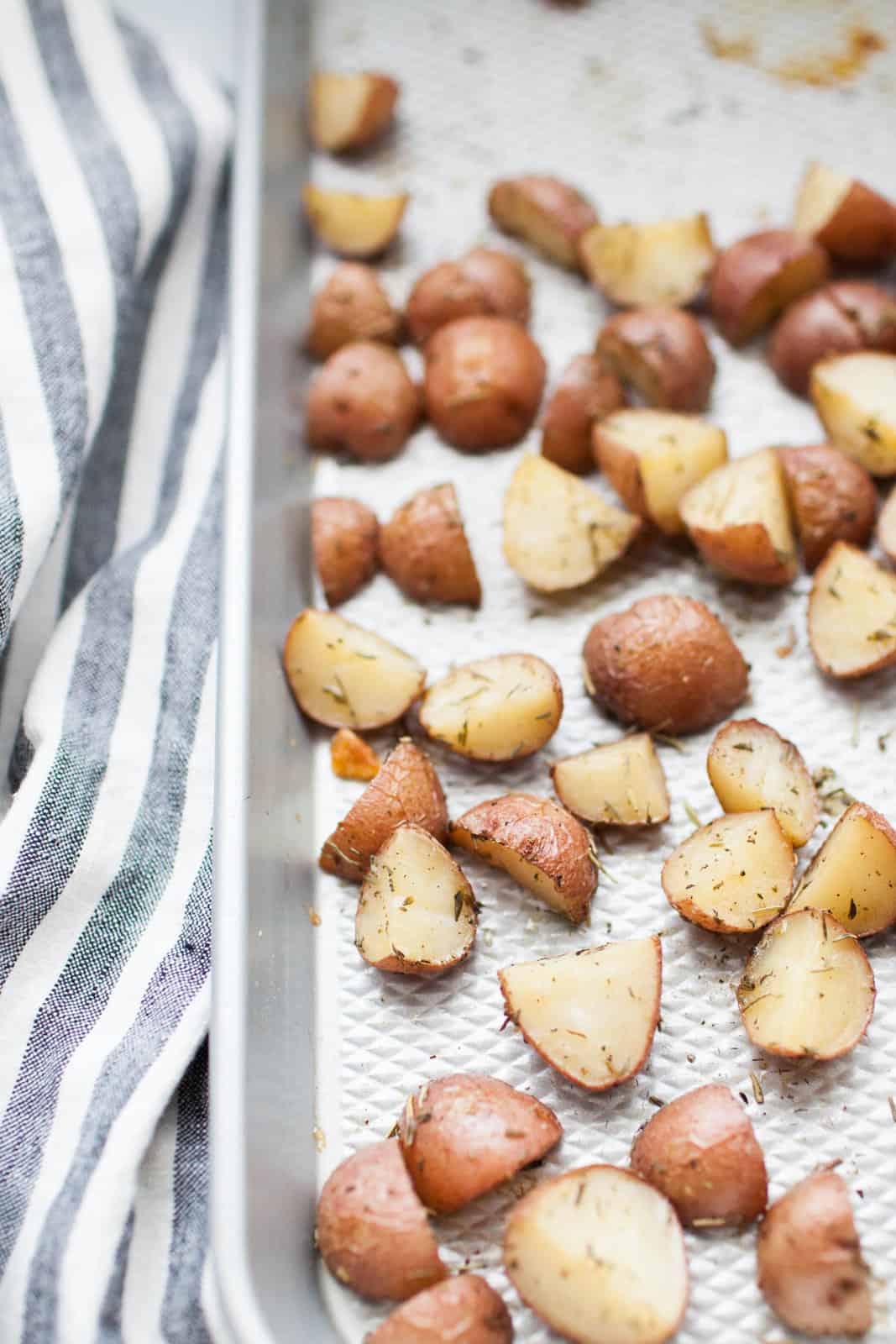 Rosemary potatoes roasted as red wedges with herbs on a baking sheet next to a striped kitchen towel.