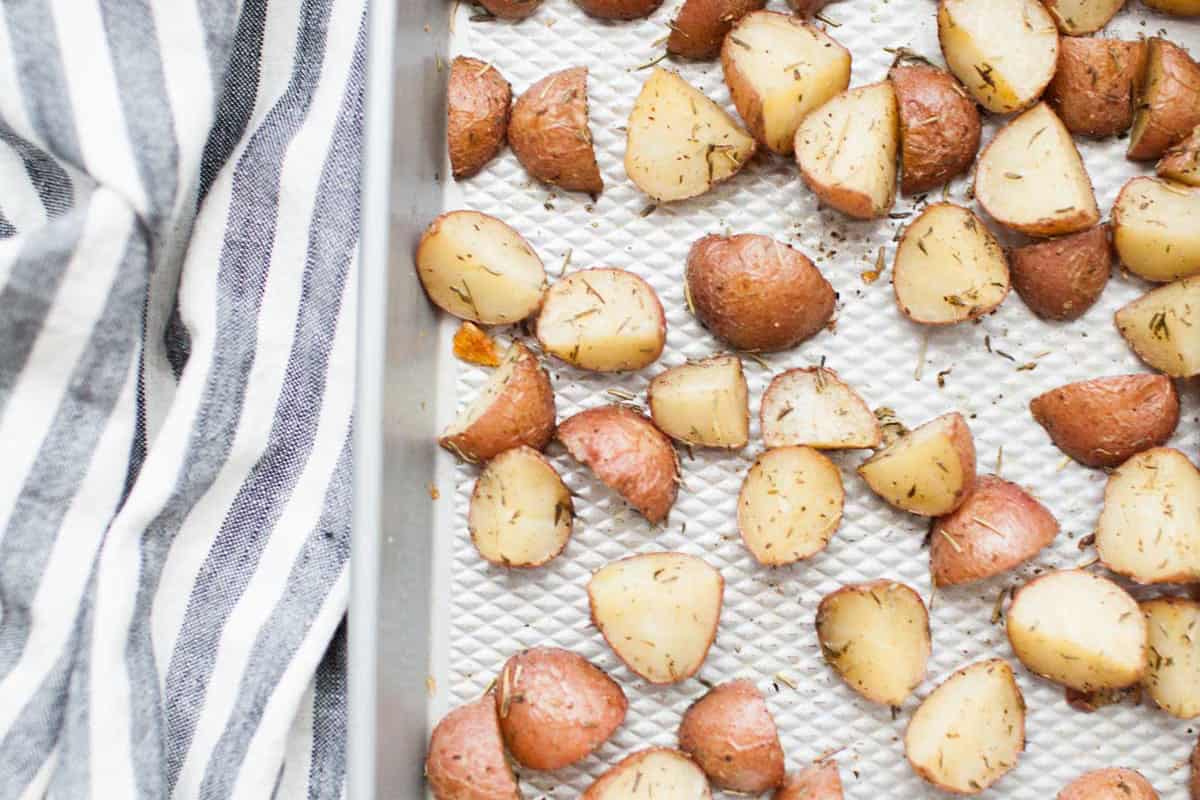 Roasted rosemary potatoes&mdash;red potato wedges with herbs&mdash;sit on a baking sheet next to a white and gray striped cloth.