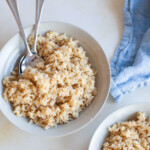 Two bowls of brown rice with two spoons in one bowl with a light blue cloth on a counter.