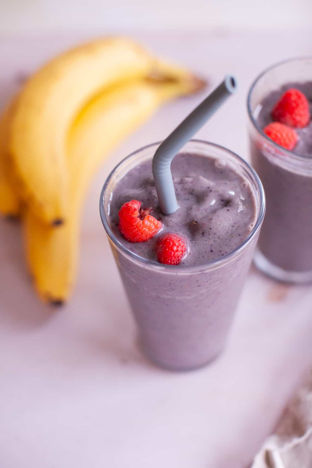 Overhead photo of two glasses with straws filled with the smoothie topped with fresh raspberries in front of two bananas.