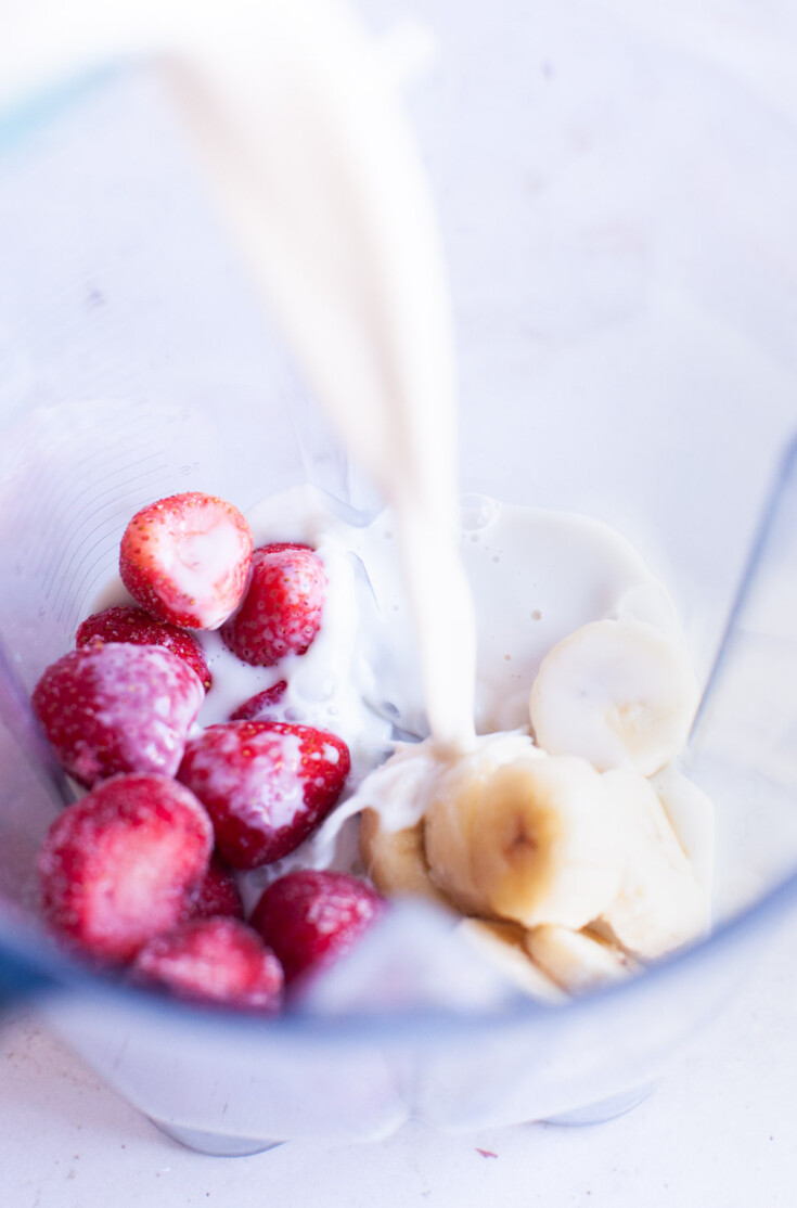 Almond milk pouring into blender on top of frozen strawberries and banana slices.