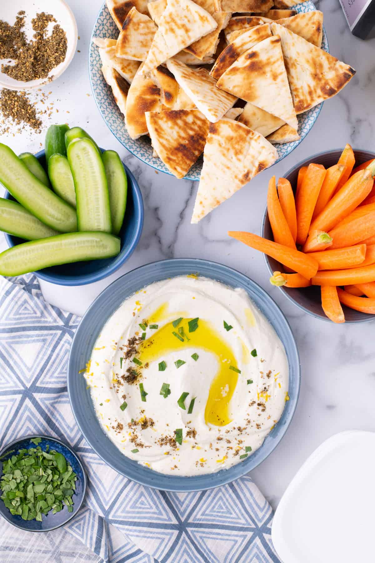 Bowls of pita bread, cucumber spears, carrot sticks, and fresh herbs around a bowl of dip.