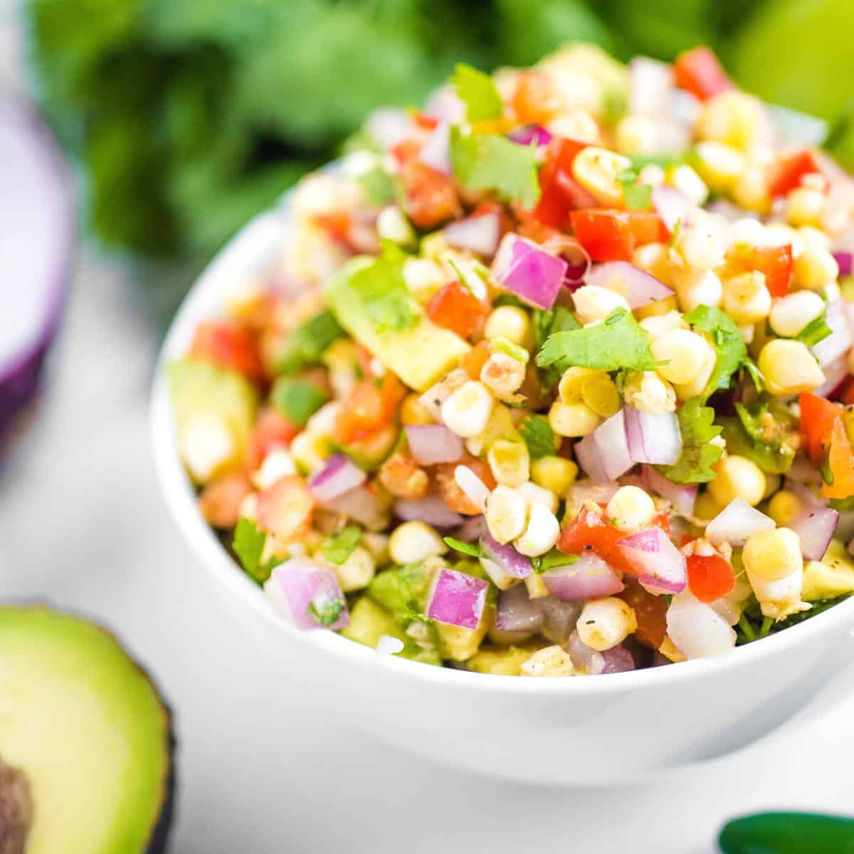 A white bowl filled with corn salsa containing corn, diced red onion, tomato, avocado, and cilantro-perfect if you're wondering what to eat with fajitas.
