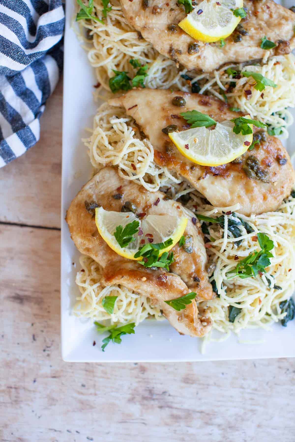 A white platter with chicken piccata and angel hair pasta with a blue and white napkin.