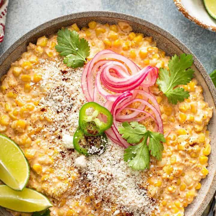 A bowl of Mexican street corn dip topped with pickled red onions, jalapeño slices, cilantro, cotija cheese, and lime wedges-perfect for what to eat with fajitas at your next fiesta.