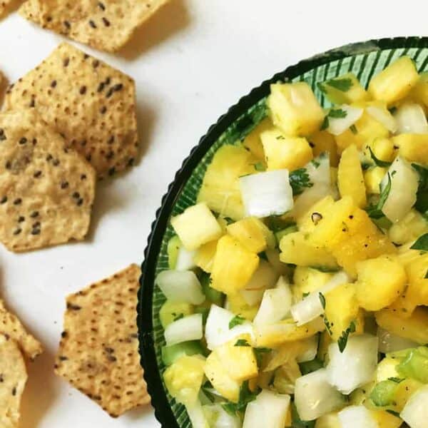 A green bowl of pineapple salsa with diced onion and herbs sits beside several hexagonal tortilla chips on a white surface-perfect for those wondering what to eat with fajitas.