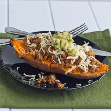 A baked sweet potato topped with cooked ground meat, shredded cheese, and guacamole sits on a black plate-a delicious twist if you're wondering what to eat with fajitas. Fork and knife are placed beside the plate.