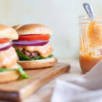 A close-up of burger sauce in a mason jar with burgers next to it.