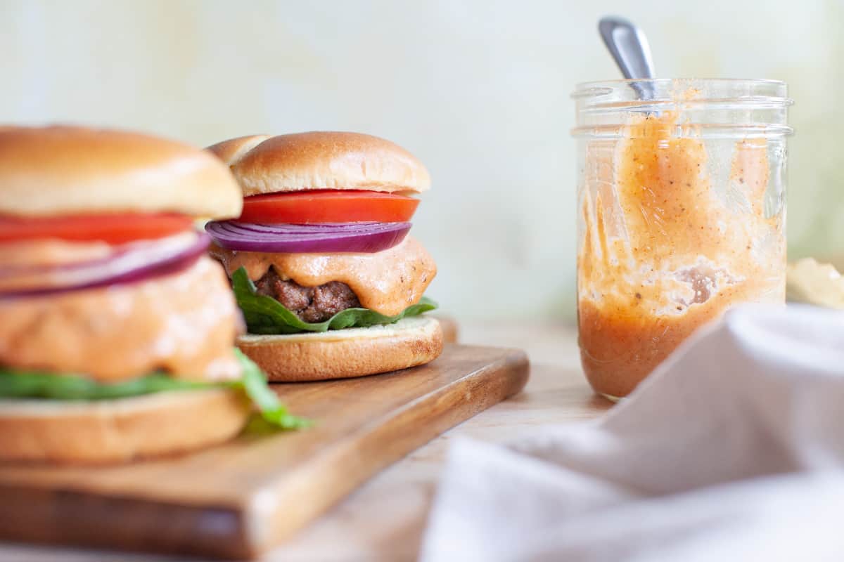 A close-up of burger sauce in a mason jar with burgers next to it.