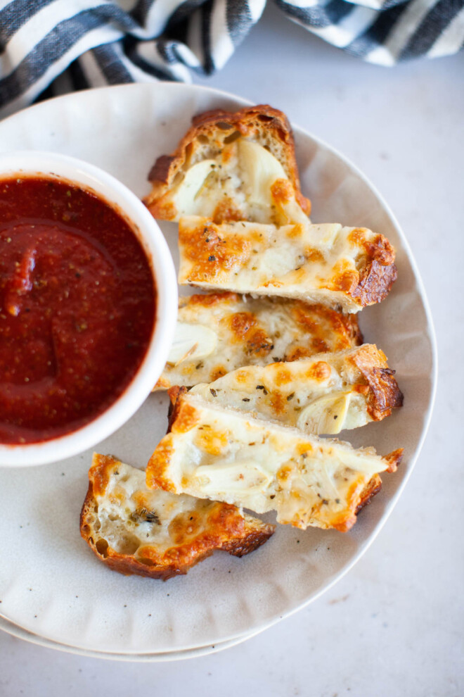 Overhead shot of thin slices of cheesy garlic bread on a small plate with a small bowl of marinara.