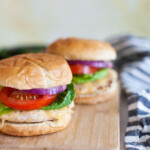 Two whole grilled chicken burgers with cheese, lettuce, tomato, red onion served with a bun on a wooden serving platter.