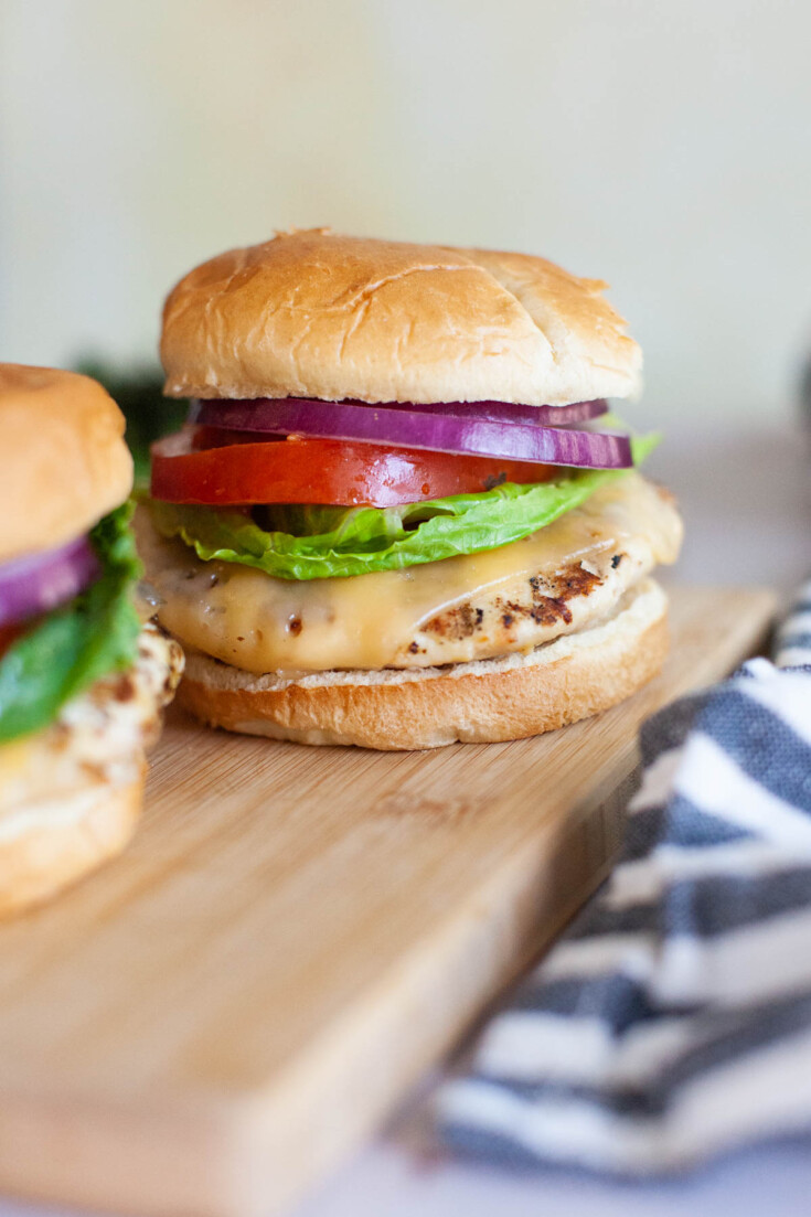 Grilled chicken burger with cheese, lettuce, tomato, red onion served with a bun on a wooden serving platter.