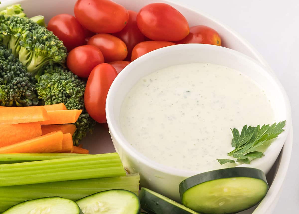 Close up photo of homemade ranch dressing in a small bowl surrounded by fresh vegetables in a larger serving bowl.