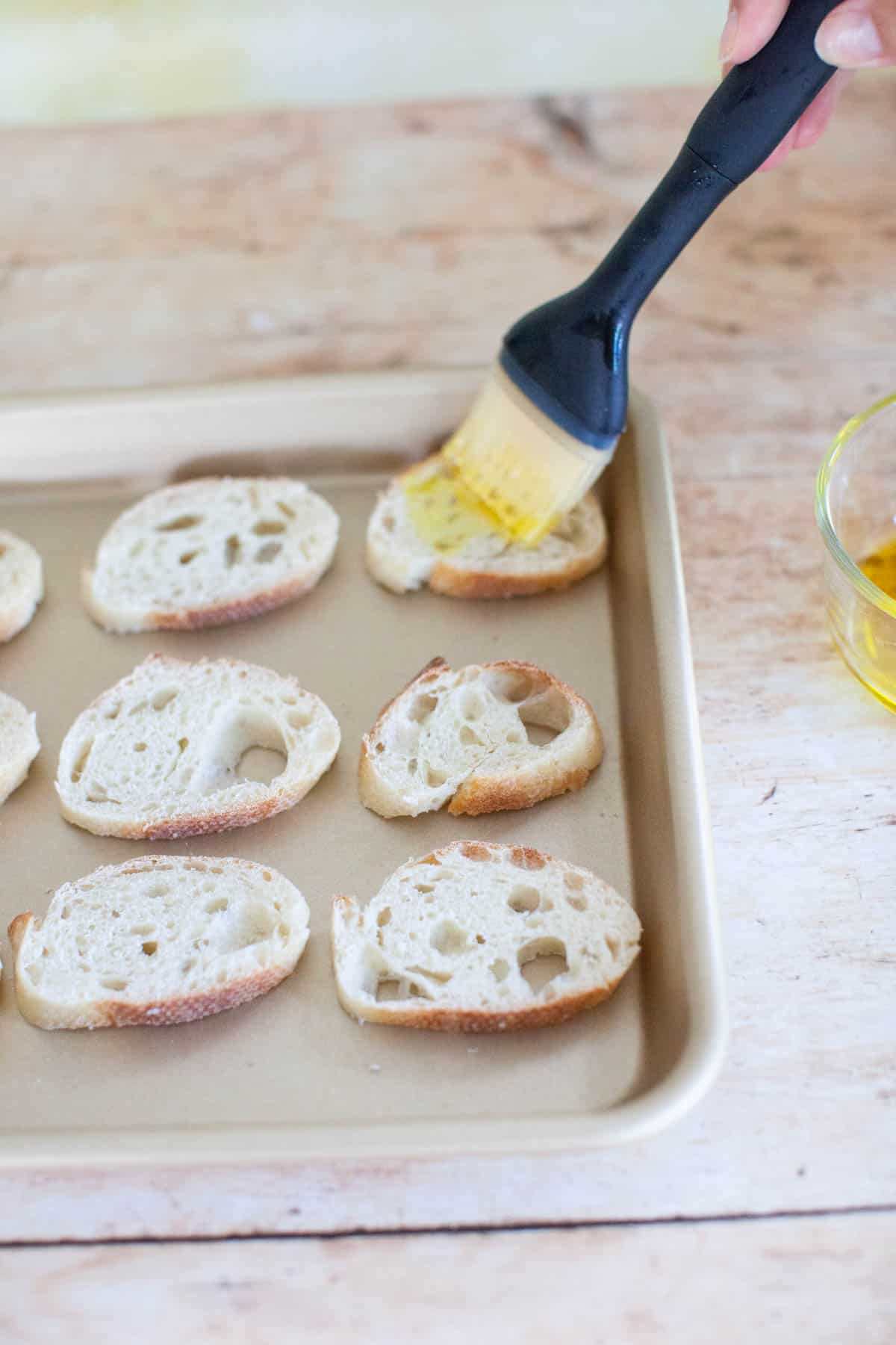 Slices of baguette on a baking sheet, being brushed in olive oil with a basting brush.