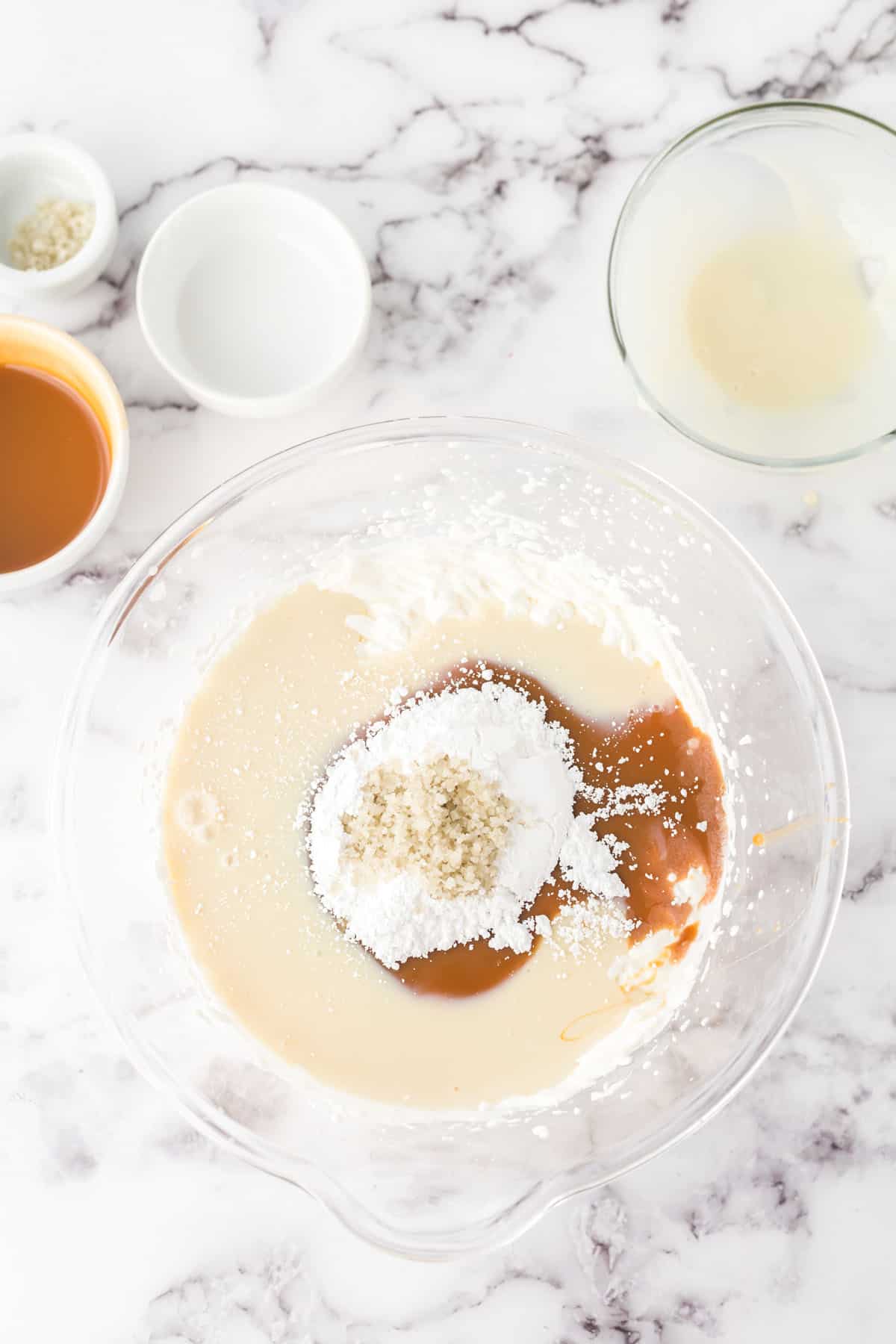 Overhead photo of sweetened condensed milk, caramel sauce, sea salt, powdered, and whipped cream in a medium-sized glass boxing bowl.