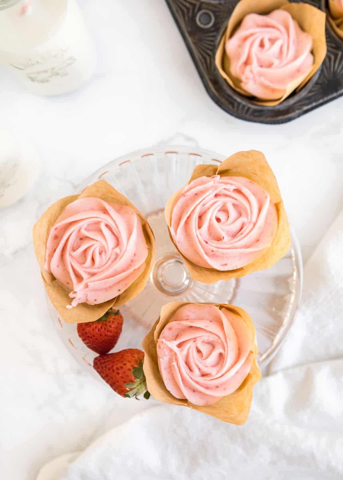 Overhead shot of three strawberry buttercream cupcakes and two whole strawberries on a small, glass cake stand. The top left and right corners are accented by a glass of milk and a cupcake in it's parchment liner in the muffin tin.