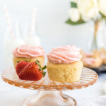 Close-up photo of two cupcakes and two whole strawberries on a small, glass cake stand with two milk jars and a small bouquet blurred in the background.