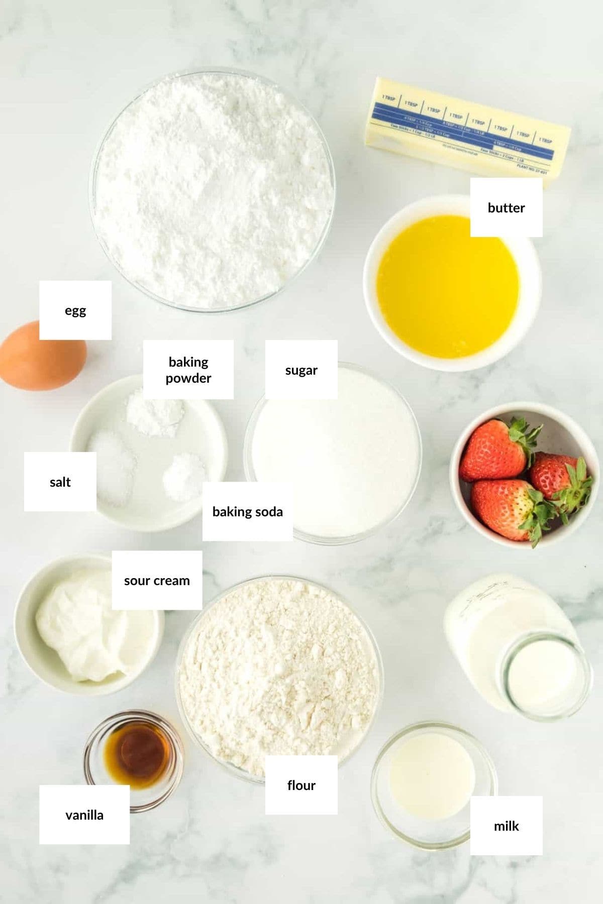 Various baking ingredients for Strawberry Vanilla Cupcakes, including flour, sugar, powdered sugar, butter, egg, milk, sour cream, vanilla, baking powder, baking soda, salt, and fresh strawberries on a white surface.