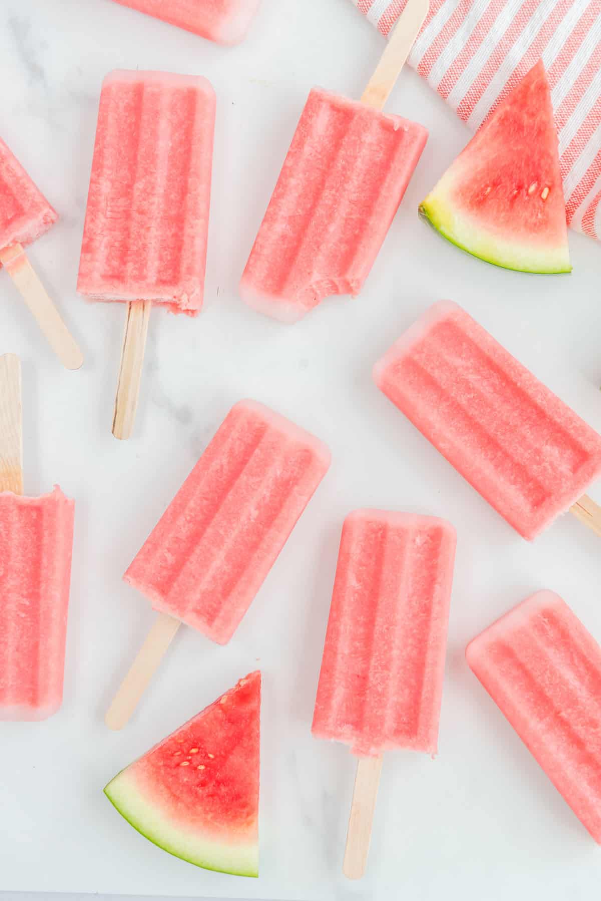 Overhead photo of multiple watermelon popsicles accented by watermelon slices in the upper righthand and lower lefthand corners. One of the popsicles has a small bite taken out of it.