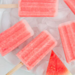 Close-up, overhead photo of a plate of ice cubes topped with watermelon popsicles stacked on top of each other. Accented by a sliced watermelon on the bottom of the photo.