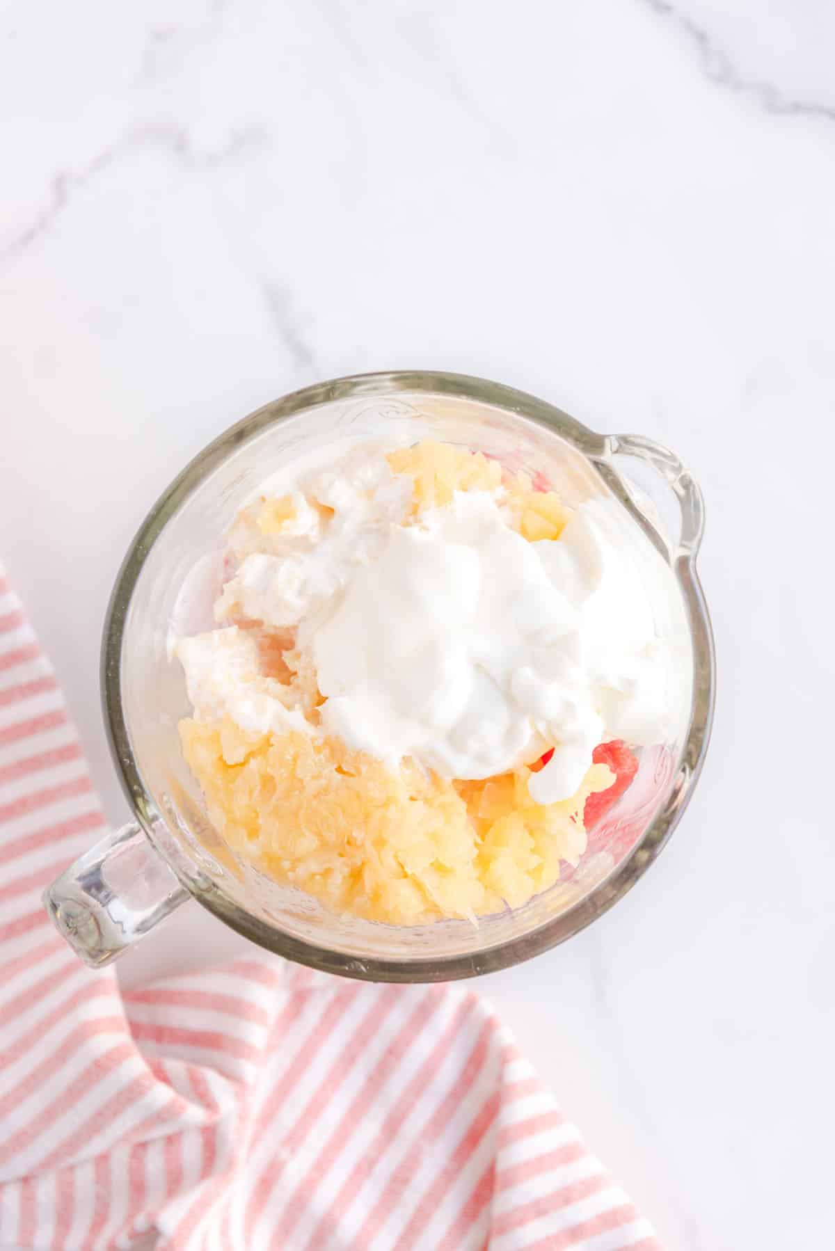 Overhead shot of blender filled with watermelon, crushed pineapple, heavy cream, lemon juice and greek yogurt. Bottom left corner accented with a pink and white-striped cloth.