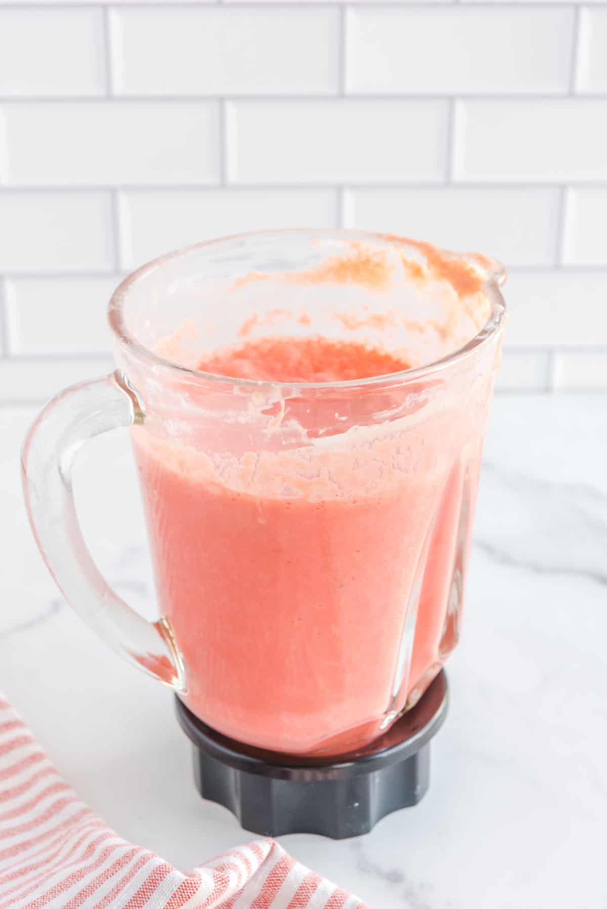 Straight on shot of blender with fully blended watermelon, crushed pineapple, heavy cream, lemon juice and greek yogurt. Bottom left corner accented with a pink and white-striped cloth.