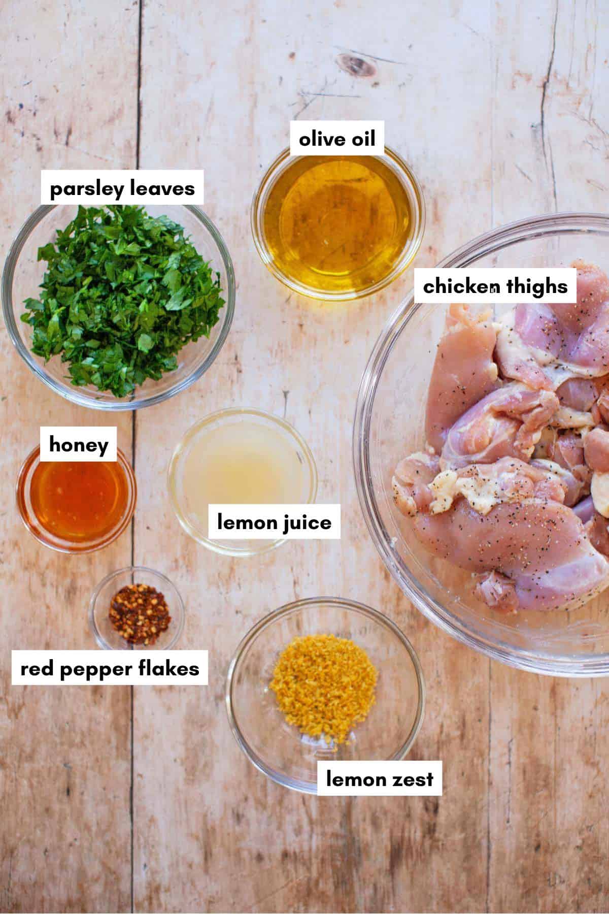 A glass bowl of raw lemon chicken thighs surrounded by small bowls of parsley, olive oil, honey, lemon juice, red pepper flakes, and lemon zest, with labels pointing to each ingredient.