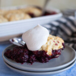 Blackberry cobbler topped with whipped cream on small, gray plate with spoons on the side.