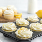 Lemon cupcakes with lemon slice garnish in a cupcake tin. Lemon cupcakes on a wooden serving platter in the background.