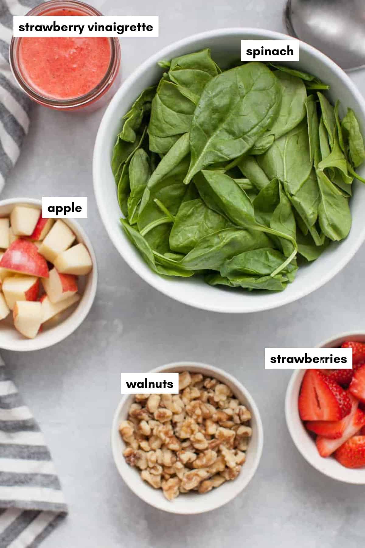 Bowls of spinach, chopped apples, walnuts, and sliced strawberries for a strawberry spinach salad, served with a jar of vinaigrette.