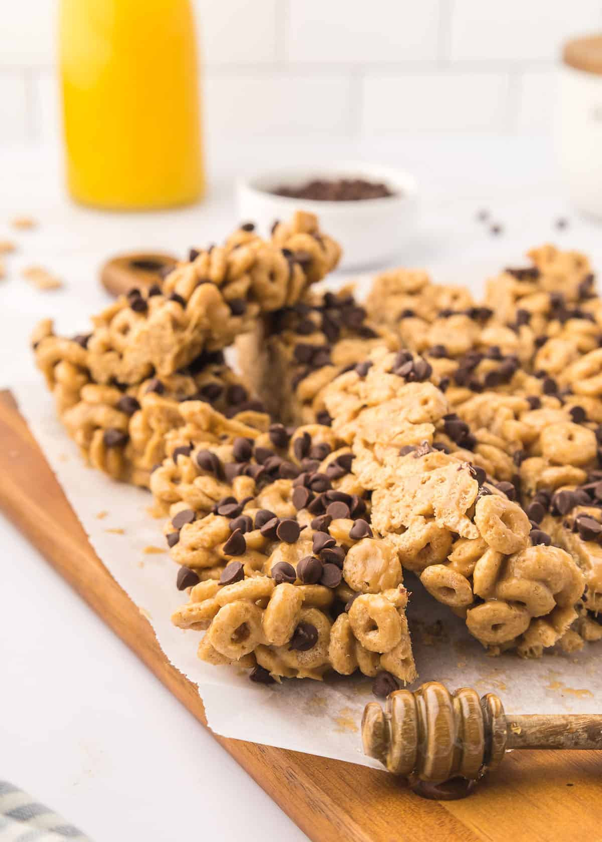 Slices of cereal bars randomly stacked on top of each other on a wooden cutting board accented with a wooden honey dipper.