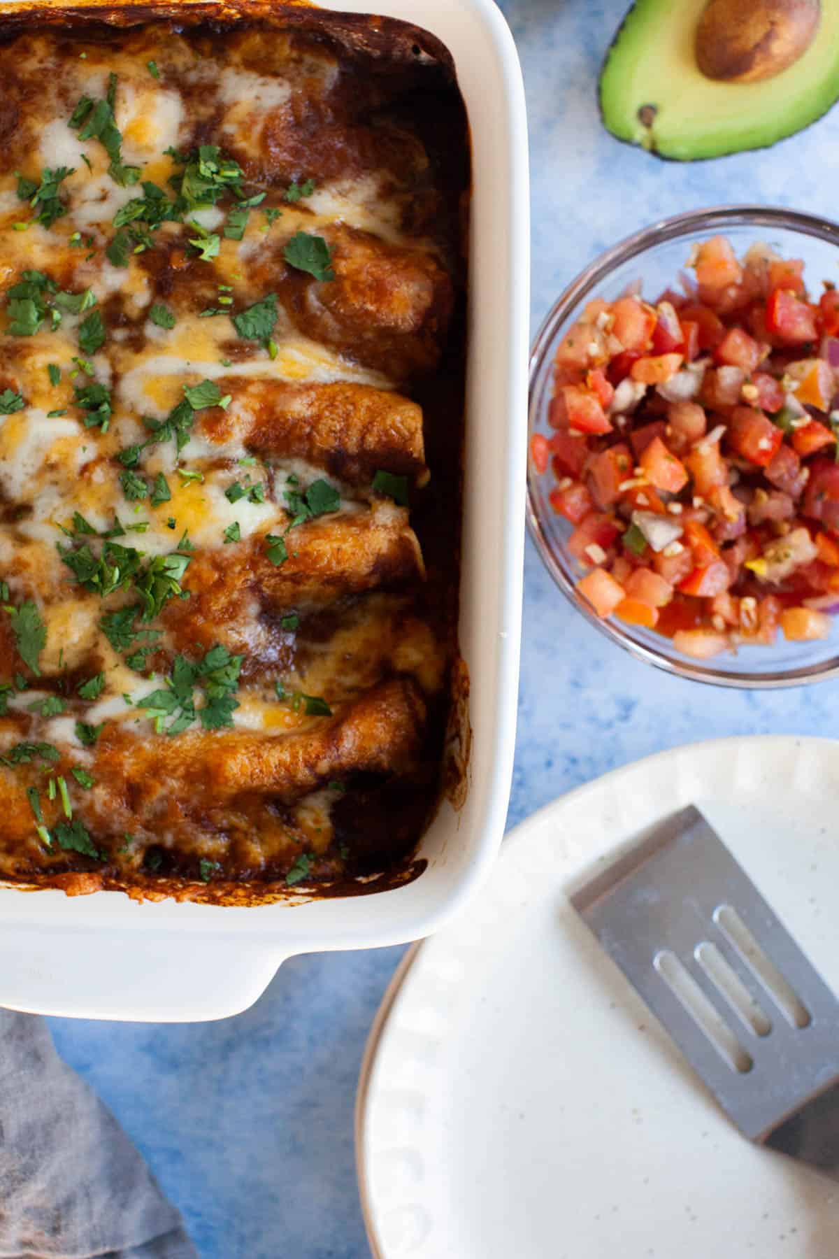 Overhead shot of fully cooked enchiladas. Accented by a bowl of pico de gallo and an avocado.