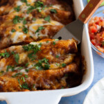 An enchilada being taking from the serving dish with a small, metal spatula.