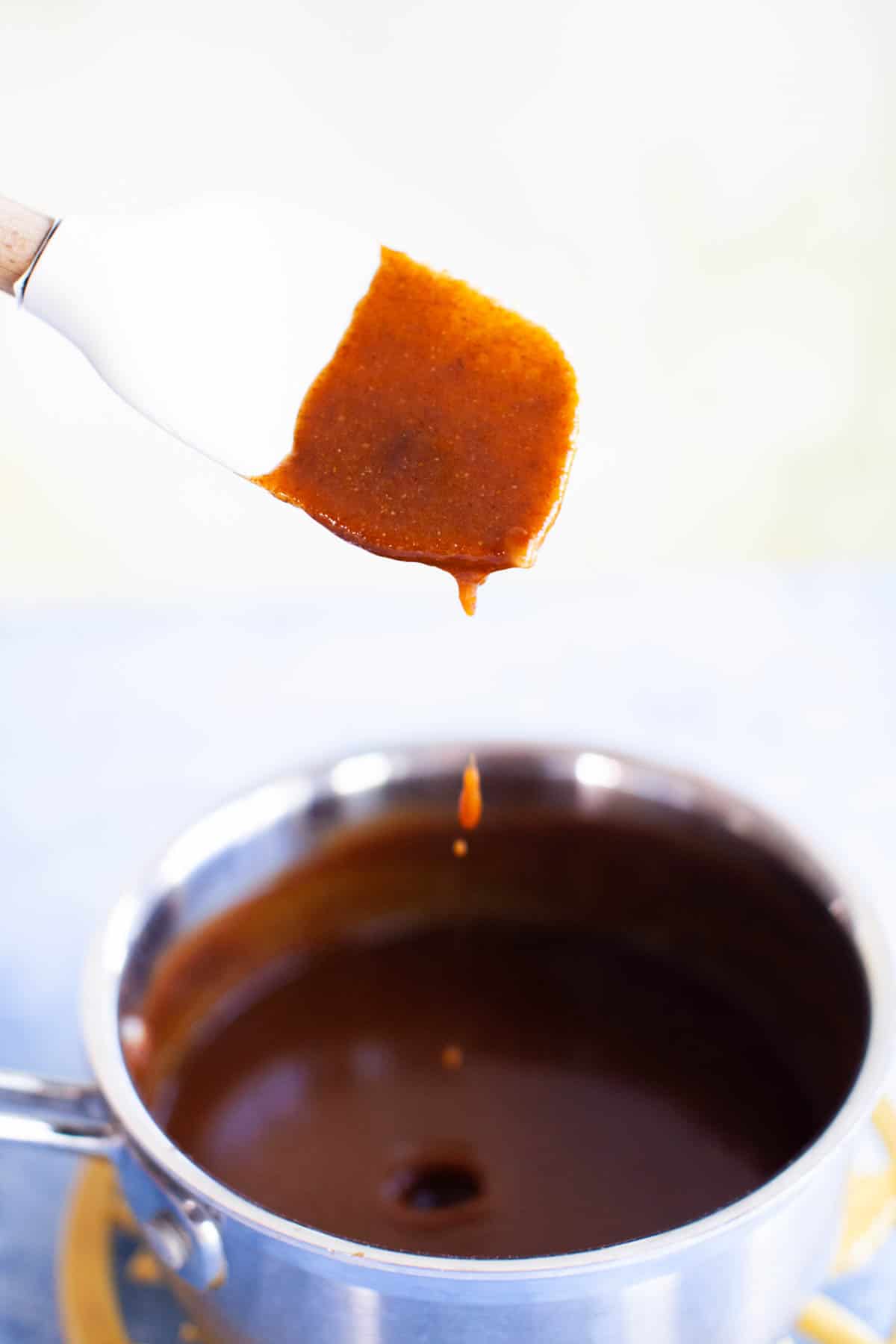 White rubber spatula being lifted out of the small pot and enchilada sauce dripping off into the pot.