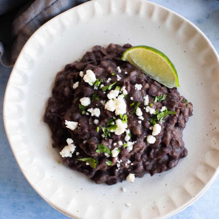 Close up refried black beans topped with cotija cheese and a lime wedge situated on a small white plate.