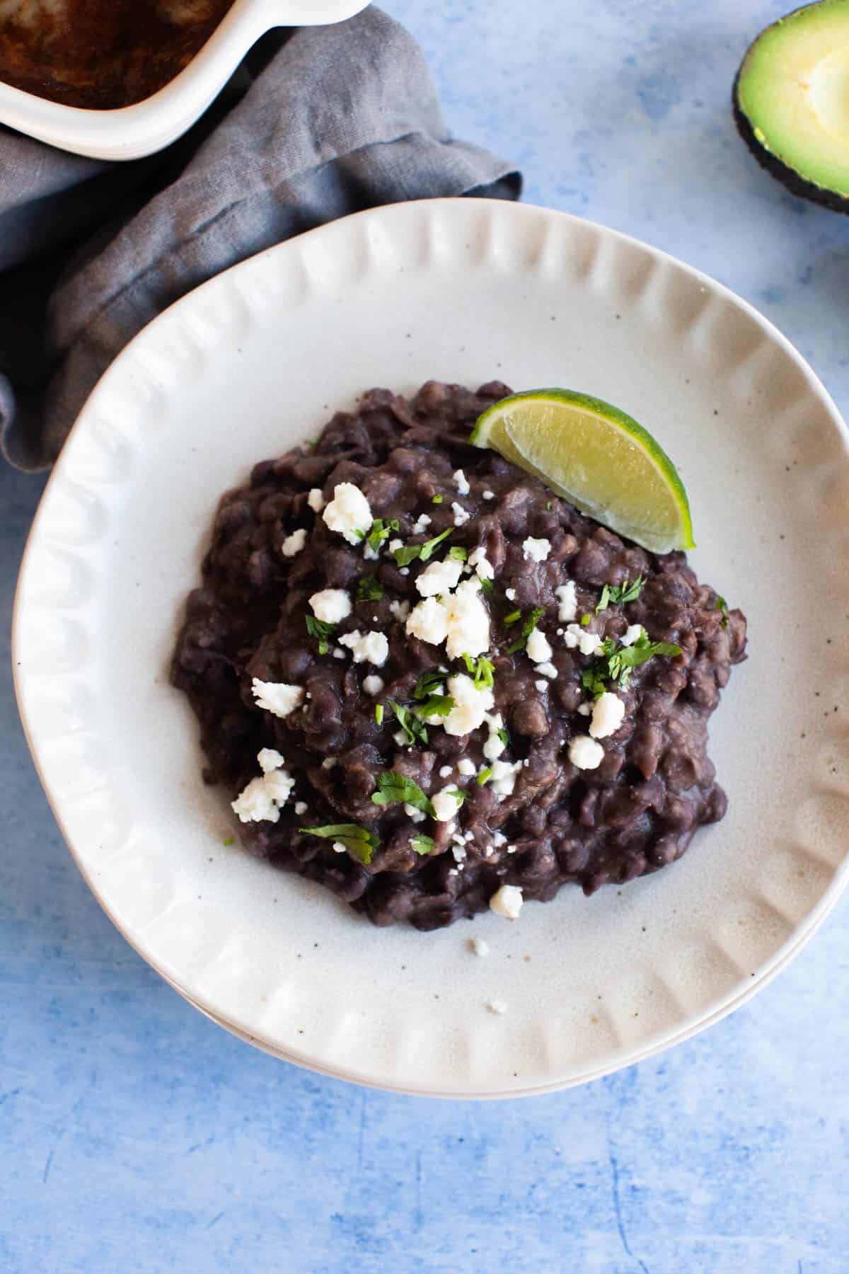 Close up refried black beans topped with cotija cheese and a lime wedge situated on a small white plate.