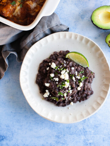 Overhead refried black beans topped with cotija cheese and a lime wedge situated on a small white plate.